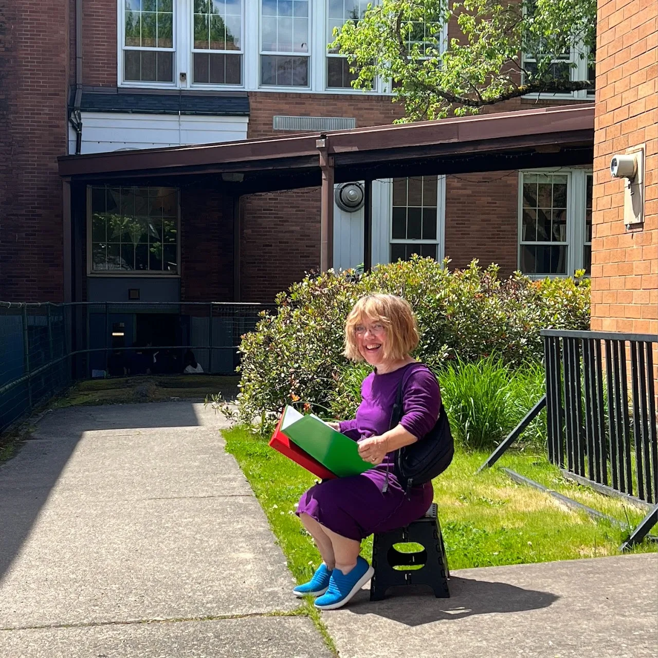 Meg Weber smiling outdoors, seated on her folding stool with a colorful folder outside a school