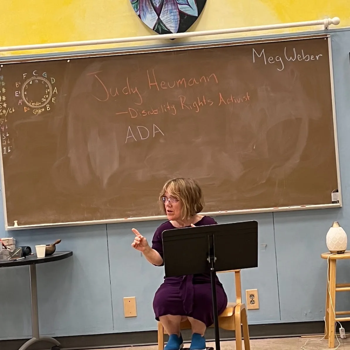 Meg seated and pointing as she teaches, with 'Judy Heumann — Disability Rights Activist' and 'ADA' written on the chalkboard behind her